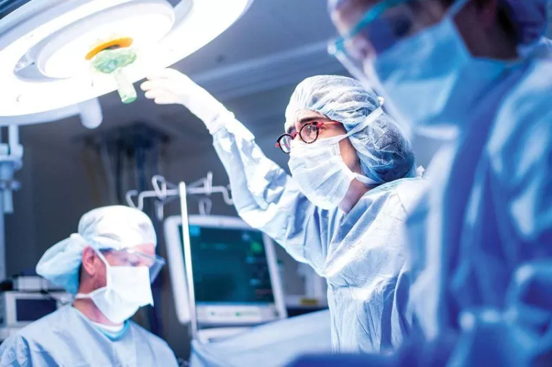 Tippi MacKenzie, in surgical scrubs, reaches gloved hand towards a light above an operating table in the operating room