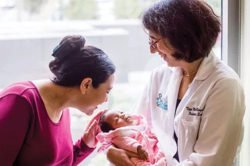 Tippi MacKenzie, woman in white lab coat, holds newborn baby, Elianna, while her mother looks on.