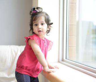 A young child with curly hair, wearing a pink top and dark pants, stands by a window ledge indoors and looks towards the camera.