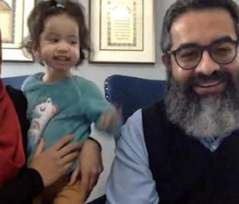 A happy family consisting of a woman wearing a red hijab, a young child in the middle, and a bearded man on the right, all smiling and posing together in front of a computer camera in a room with wall decorations.