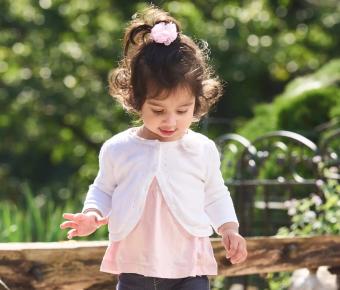 A young child with curly hair, wearing a pink top and dark pants, stands by a window ledge indoors and looks towards the camera.
