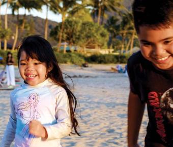 Two children are running on a beach during sunset, with palm trees and other beachgoers in the background.