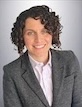 Dawn Gano smiles in dark blazer with white shirt, short curly hair, against a grey backdrop