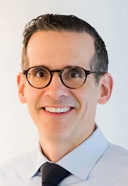 Juan Gonzalez Velez, MD, MS, PhD, smiling with blue dress shirt tie and grey background